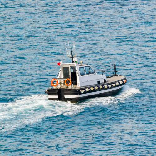 A white pilot boat with a black hull speeds across a blue ocean. The boat has a small cabin, a flag flying from the mast, and a life ring hanging on the side.